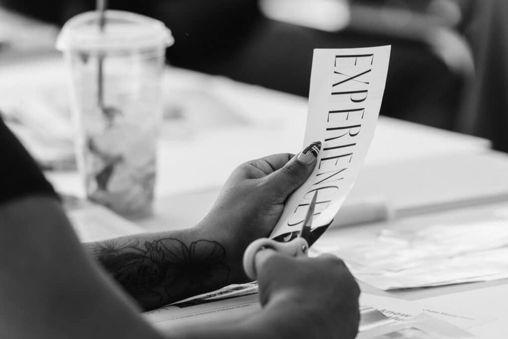 Black-and-white photograph of Black woman's hands with scissors, cutting out the word EXPERIENCE from a piece of paper.