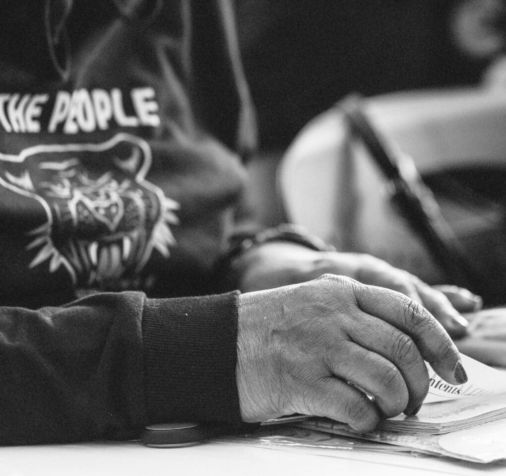 Black-and-white photograph of a Black woman at a table; her sweatshirt reads: THE PEOPLE.