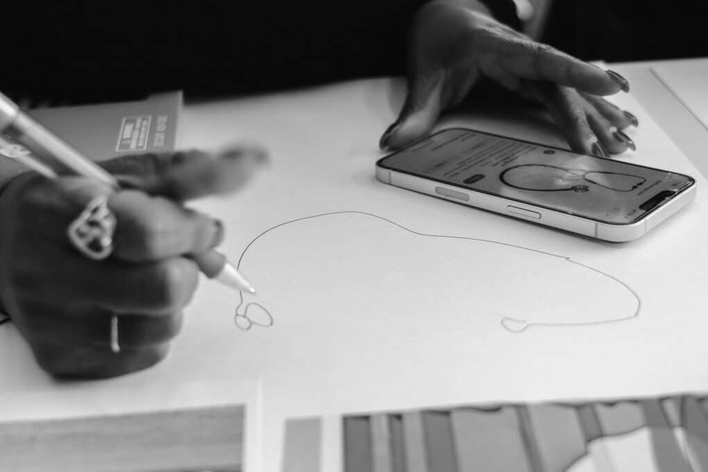 Black-and-white photograph of a young Black woman's hands drawing a stethoscope from an image on an iPhone.