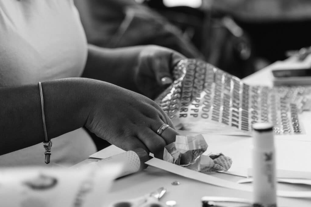 Black-and-white photograph of a Black woman's hands working with a sheet of decal letters at a table strewn with papers and books.