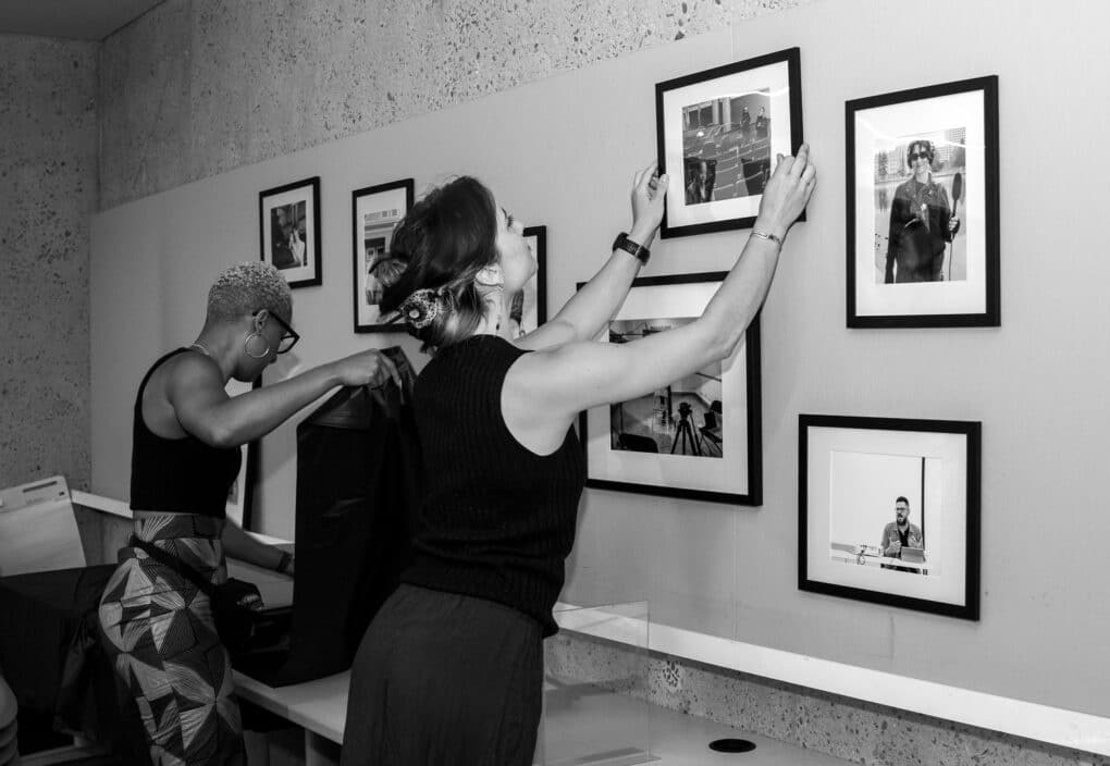 Two young women, one Black and one white, hang framed photographs on a white wall.