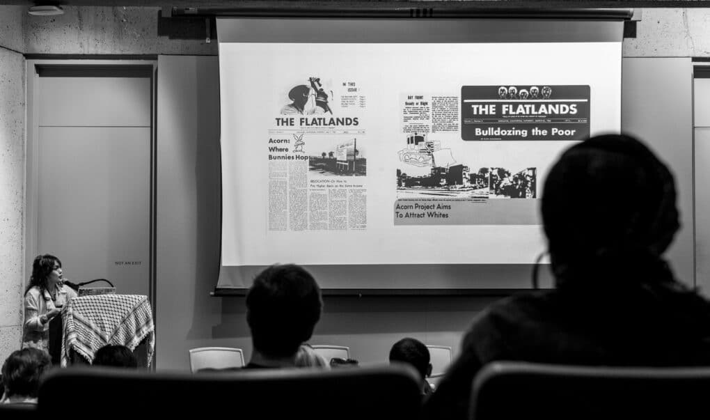 Black-and-white photograph of stage in a small auditorium, taken from the audience. On the screen are two images from "The Flatlands" newspaper, one with the headline "Bulldozing the Poor."
