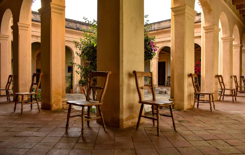 Colonnaded courtyard with trees, tile floor and wooden chairs ranged around the pillars.