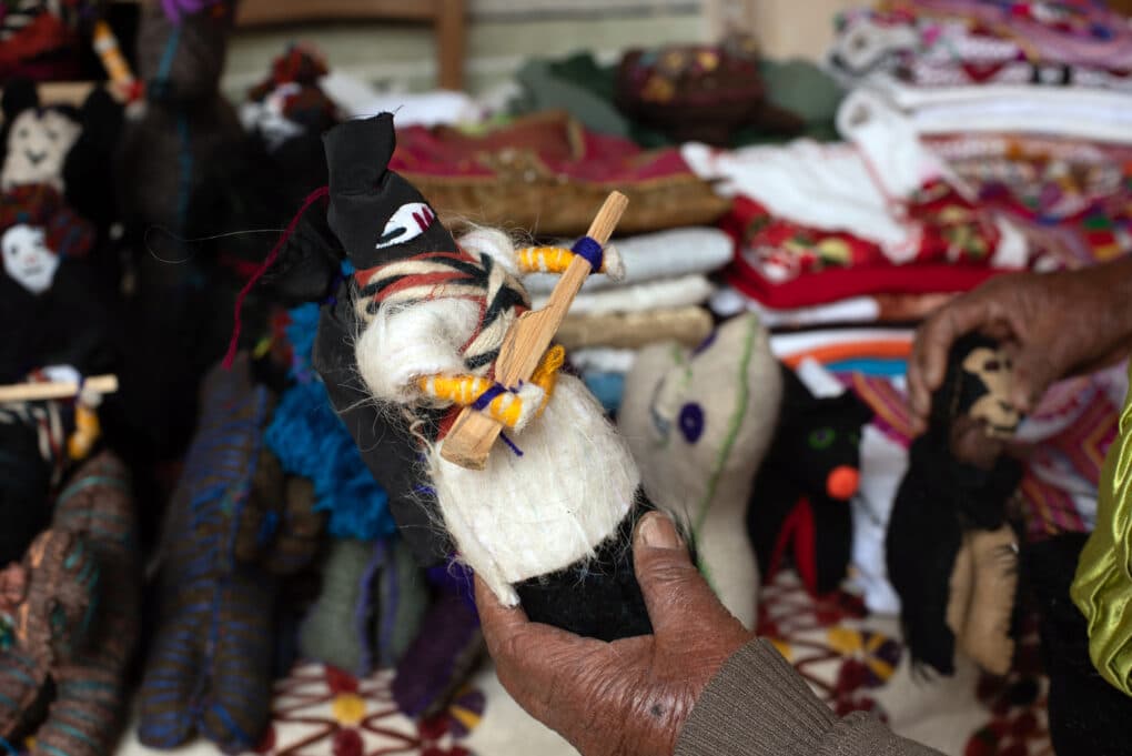 Color photograph of woolen doll held in a dark-skinned, elderly hand; in the background are piles of textiles and other woolen dolls.