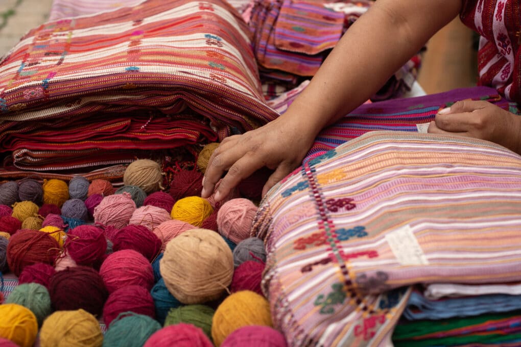 Color photograph of balls of yarn and colorful weavings, with a young person's brown-skinned hand and arm.