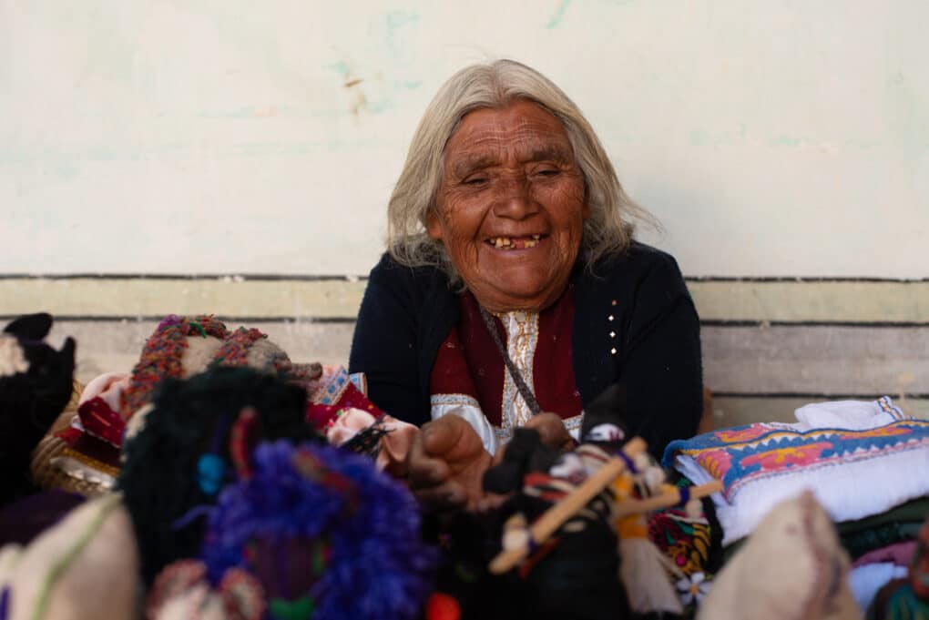 Color photograph of white-haired, dark-skinned woman smiling, photographed against a white wall. In the foreground are hand-made yarn dolls.
