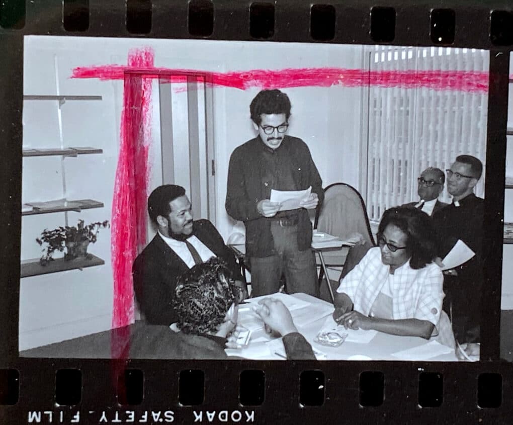 Black-and-white image from photographic contact sheet, marked with red crop mark, showing five Black men and one Black woman, seated in an office. Two of the men wear clerical collars.