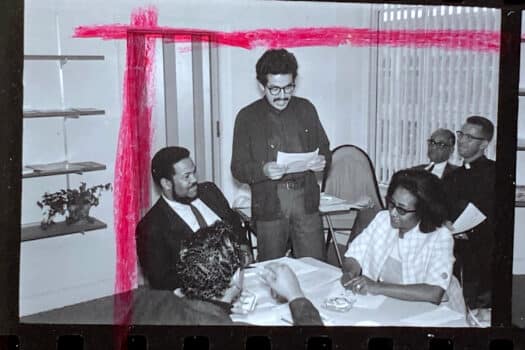 Black-and-white image from photographic contact sheet, marked with red crop mark, showing five Black men and one Black woman, seated in an office. Two of the men wear clerical collars.