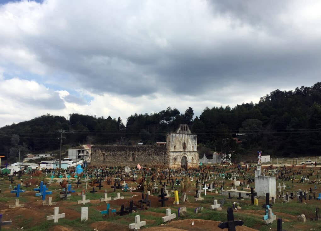 Color photograph of small stone church surrounded by crosses, under a cloudy sky with densely forested hills in the background.