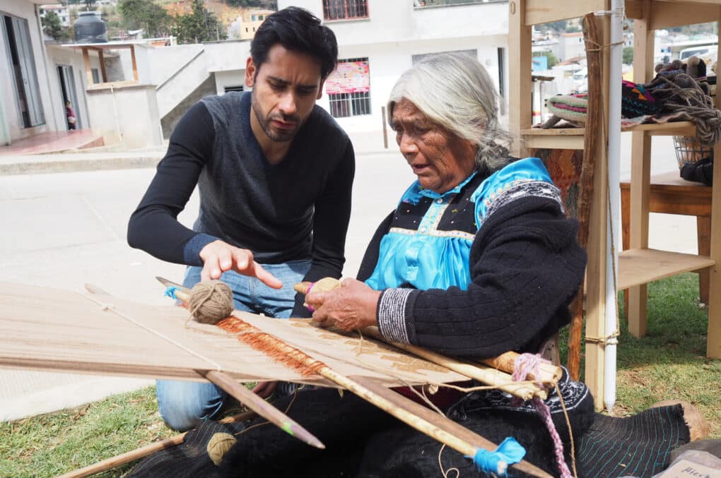 Color photograph of young man and white-haired woman at a backstrap loom, outdoors.
