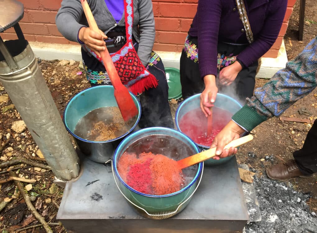 Color photograph of three women at a small outdoor stove, each stirring a bucket of dye in which floats a skein of wool (two red, one yellow) with a wooden paddle.