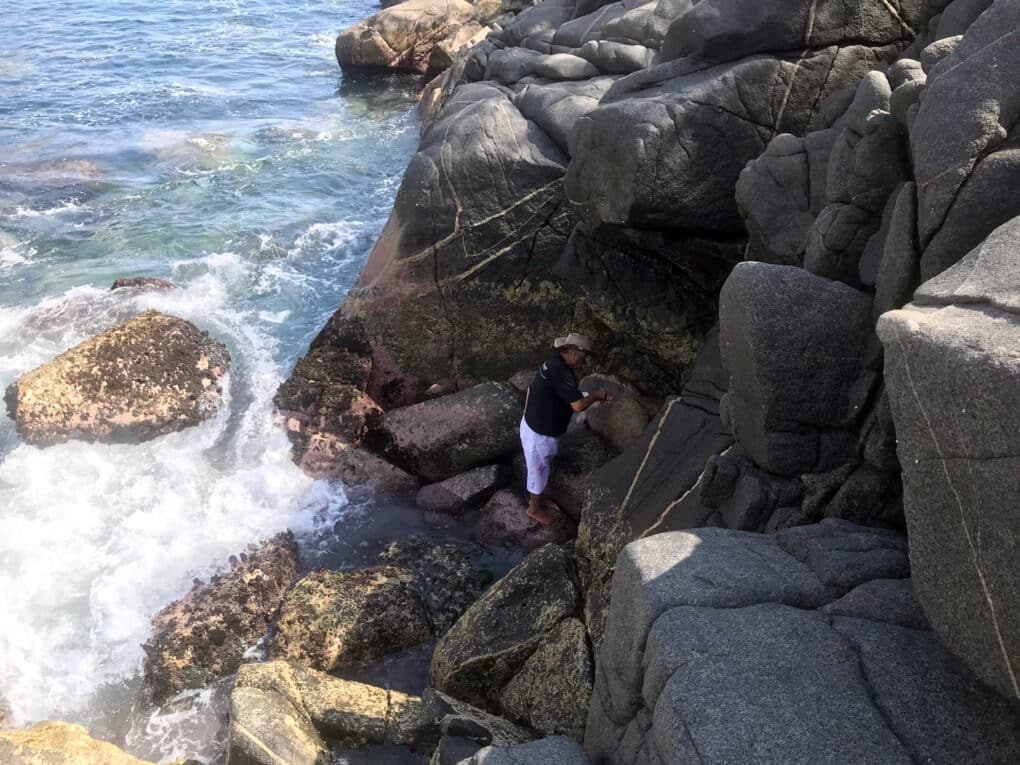 Color photo looking down from rocky seaside cliff at a man in white trousers and cowboy hat on rocks at water's edge.