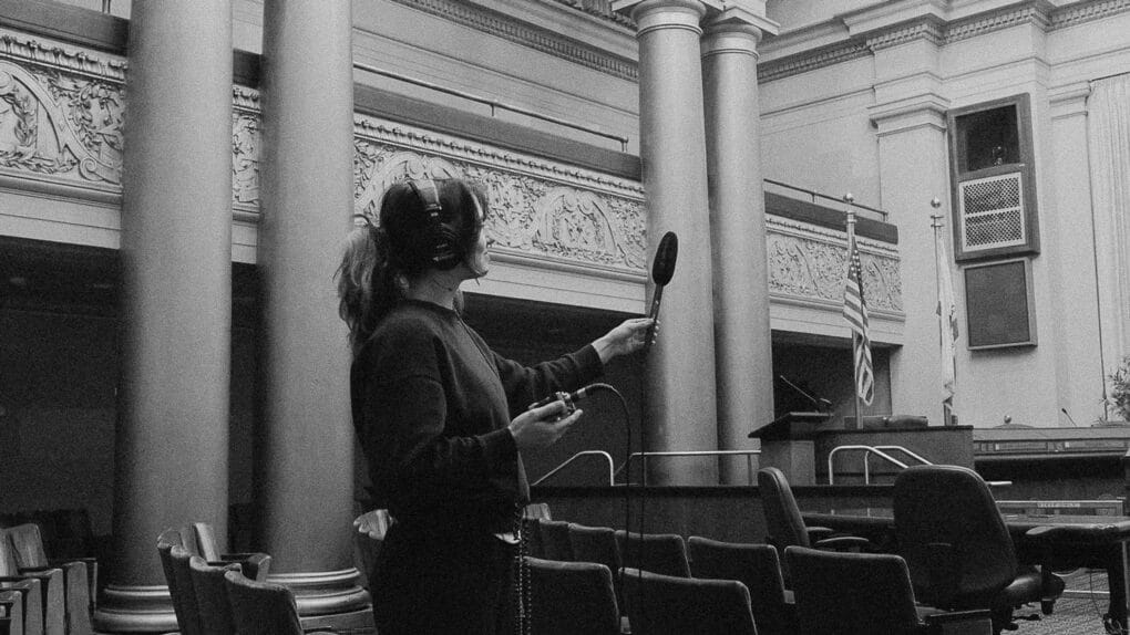 Black-and-white photograph of young white woman with headphones and microphone recording in a large ornate public chamber.