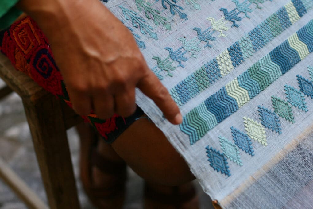 Color photograph of blue and yellow weaving, with a young woman's brown hand pointing to a detail.