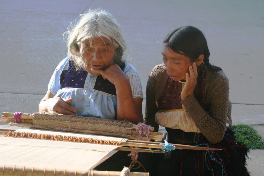 Color photograph of two women, one with white hair and the other with dark hair in braids, looking at a weaving on a backstrap loom.