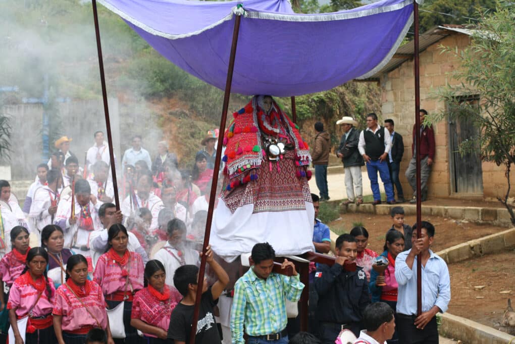 Color photograph of procession with men carrying a richly dressed statue of the Virgin under a purple fringed canopy.