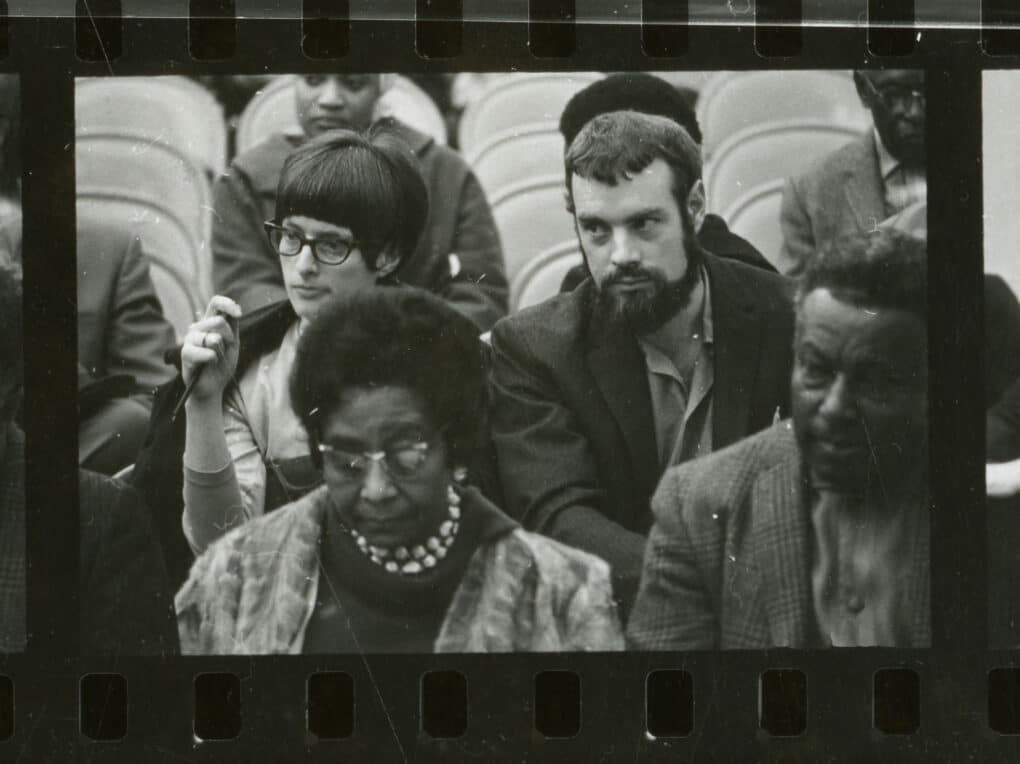 Black-and-white contact-sheet image of eight people seated in folding chairs, including a young white woman with dark short hair, bangs, and black-framed glasses.