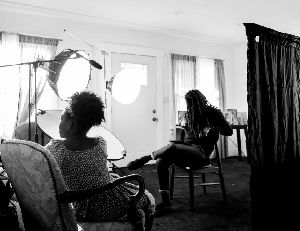 Black-and-white photograph of young Black man and Black girl in a domestic interior, wiht large photographer's lights.