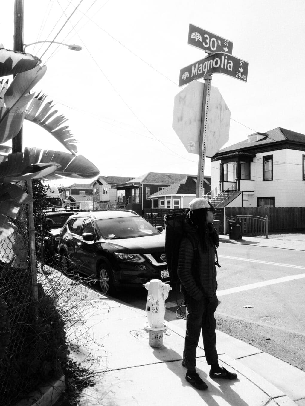 Black-and-white photograph of young black man on a street corner, with palms and single-family houses, beneath a pair of street signs reading "30th" and "Magnolia."