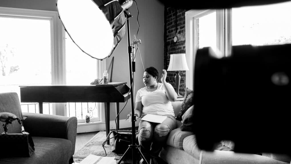 Black-and-white photograph of a young Black woman sitting beside a piano in a living room, with camera equipment all around her.