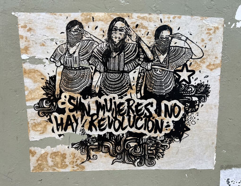 Color photograph of black-and-white poster showing line drawing of three women in Mayan dress, each masked and saluting, with a caption reading SIN MUJERES NO HAY REVOLUCION.
