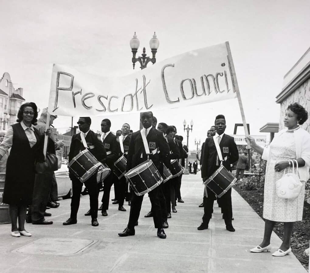 Black-and-white photograph of young Black men in drum corps uniforms, with drums, standing on a sidewalk under a banner that reads "Prescott Council." Two well-dressed middle-aged Black women hold the banner.