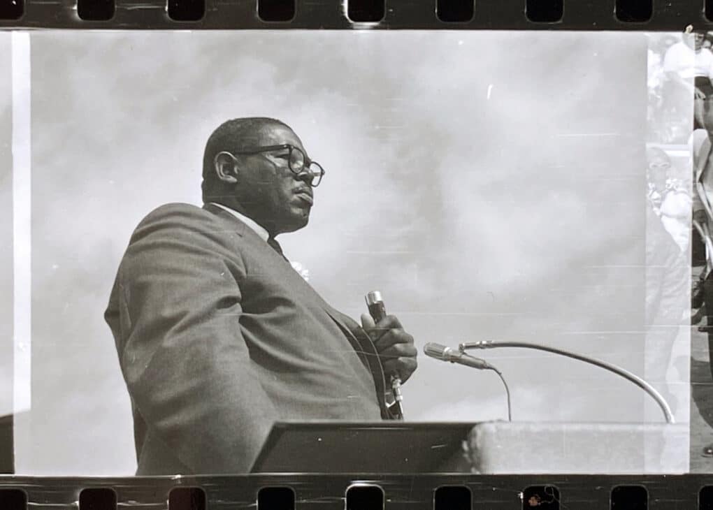 Black-and-white photograph of tall Black man in a suit and tie, standing at a podium with a microphone in one hand.