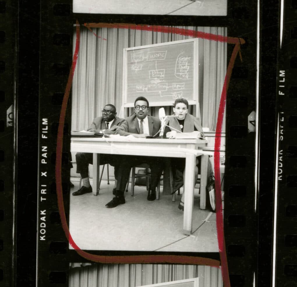 Black-and-white image from contact sheet of two men and a woman seated at a small table; behind them is a chalkboard with a diagram headed "WOPC."