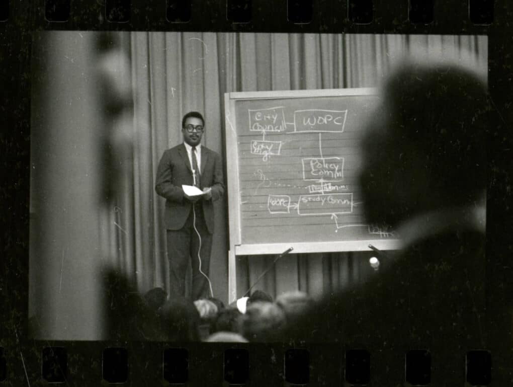 Black-and-white image from contact sheet of Black man in suit and tie addressing a group, standing beside a blackboard on which is written a diagram headed "WOPC."
