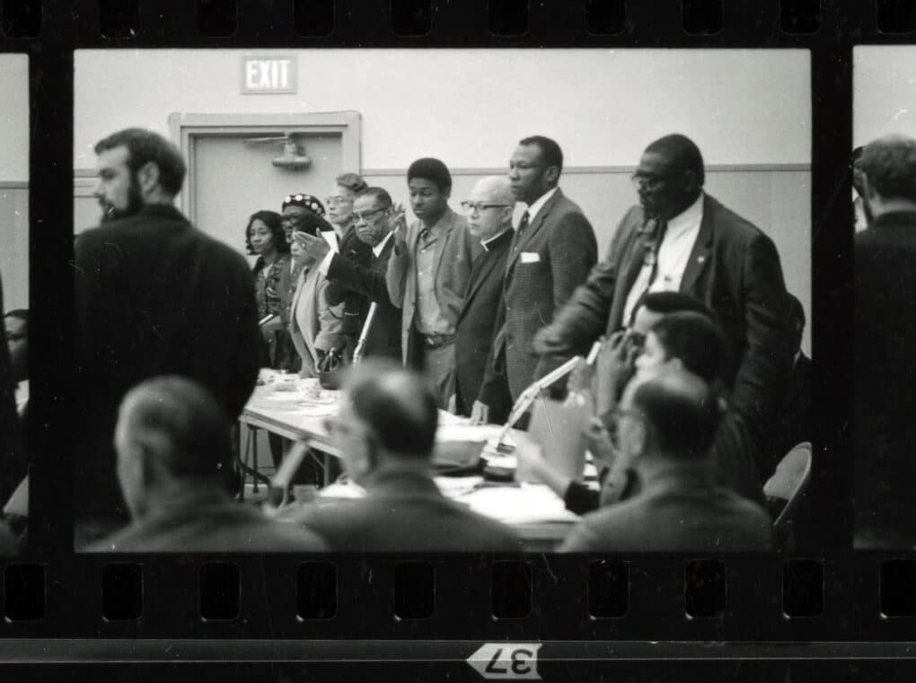 Black-and-white contact-sheet image of a group of well-dressed Black citizens standing at a large table littered with papers and microphones.