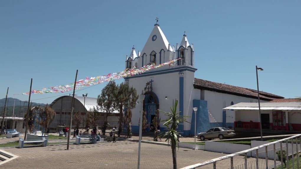 Color photograph of white stone church with flags flying from it, in front of a small paved plaza with blue sky overhead.