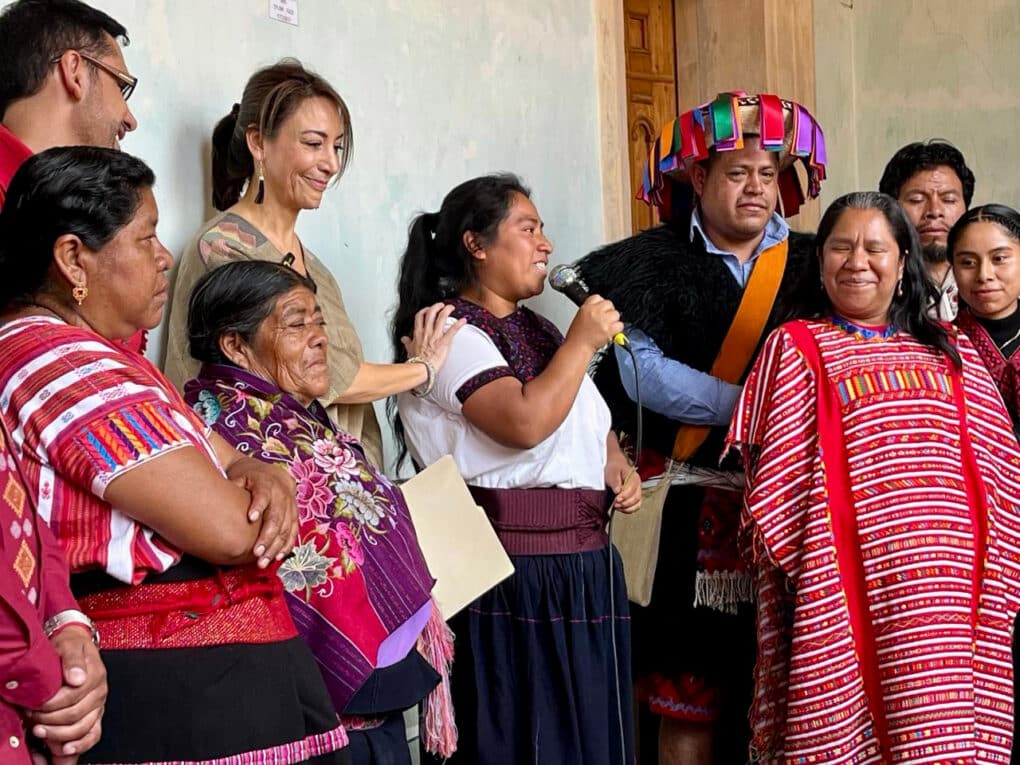 Color photograph of a small group of people, most in Mayan huipiles, gathered around a young dark-haired woman speaking into a microphone.