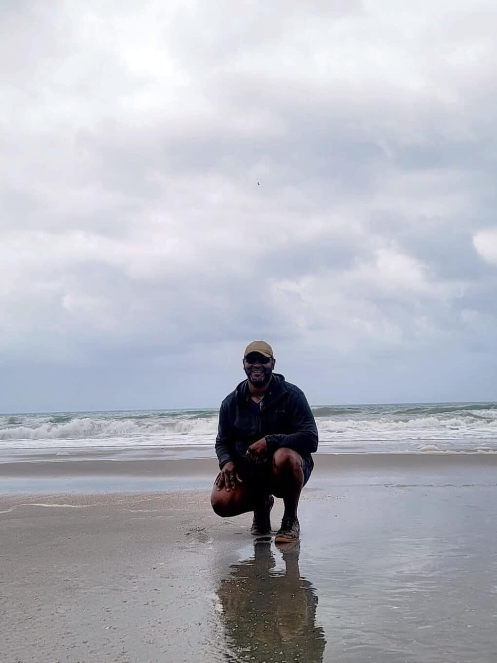 Color photograph of Black man in shorts, rain jacket, and baseball cap, squatting low on wet sand in front of the ocean on a cloudy day.