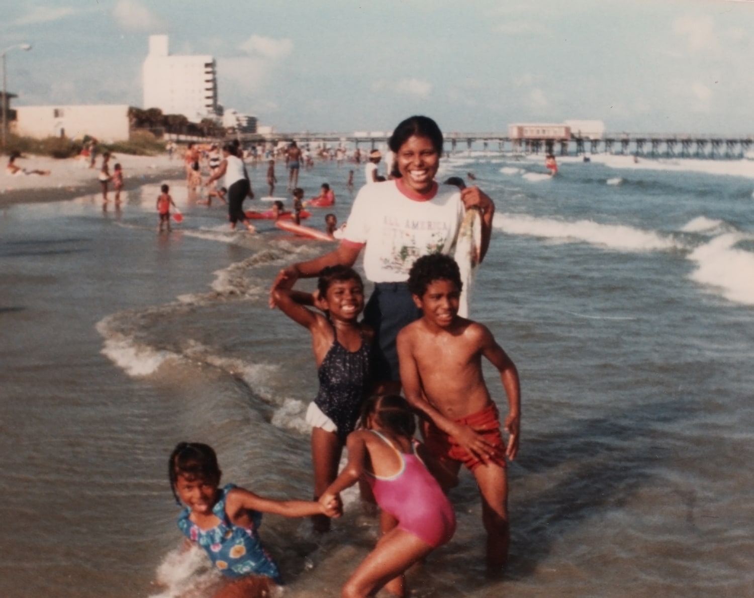 Click to Enlarge Color photograph of young Black woman and four small Black children playing in the waves and smiling for the camera.