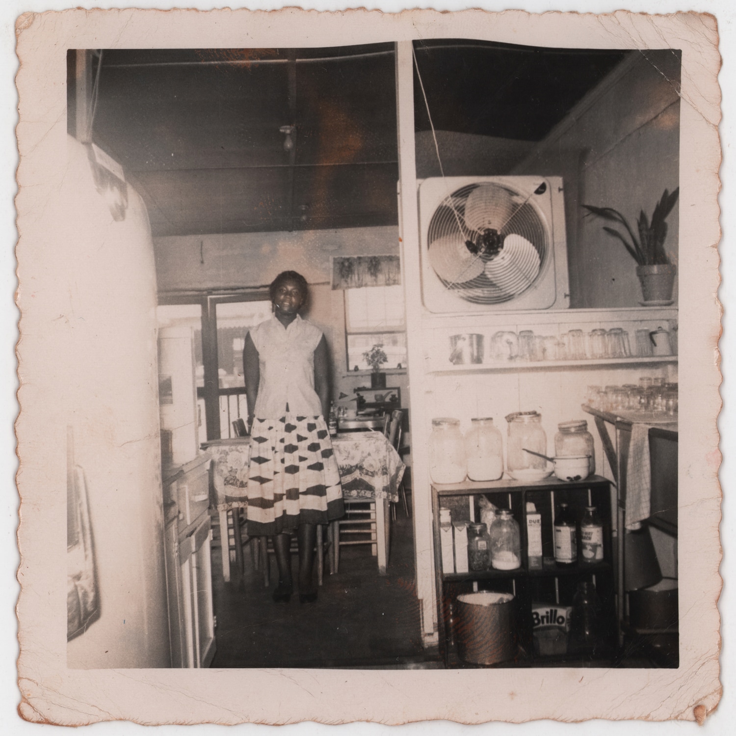 Click to Enlarge Black-and-white photograph of young Black woman in small interior with table, chairs, refrigerator, and fan.