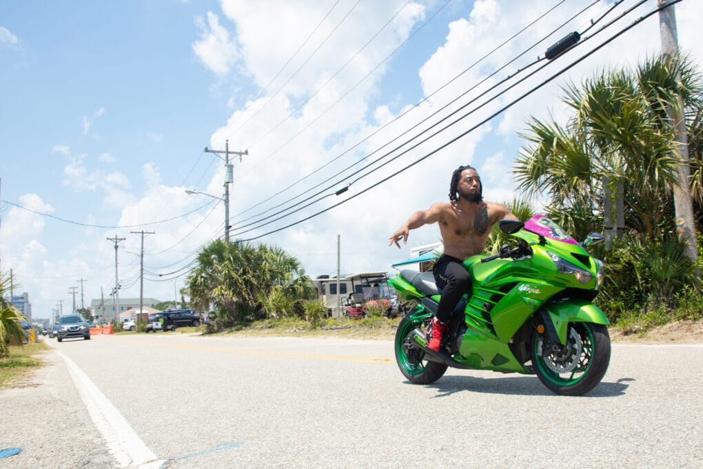 Color photograph of young Black man, shirtless, riding a green motorcycle along the highway outside a small beach town.