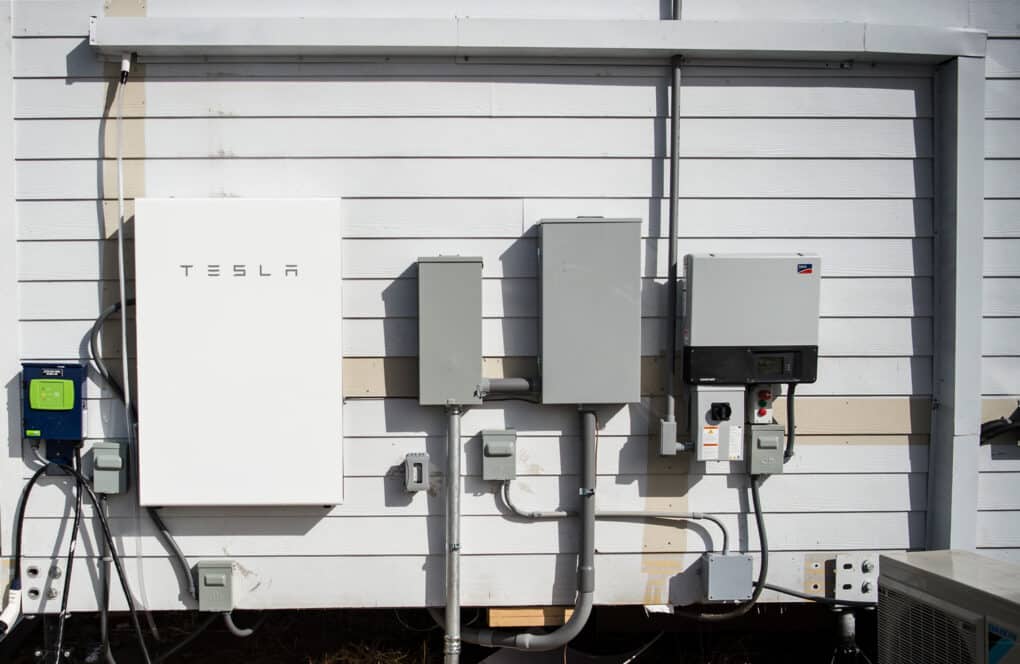 Color photograph of Tesla home battery and other electrical boxes on the wall of a white clapboard house.