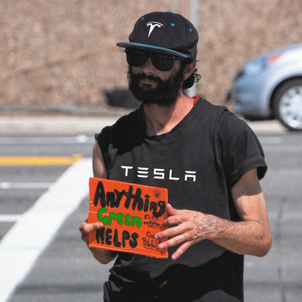 Color photograph of young white man wearing sunglasses, black Tesla baseball cap and black Tesla t-shirt, holding small cardboard sign reading "Anything Green Helps."