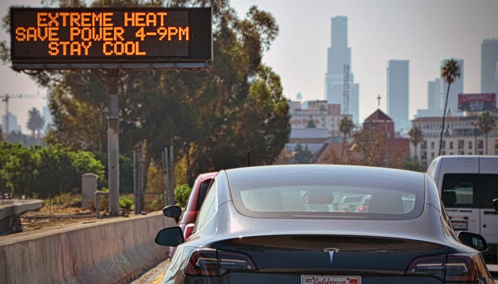 Color photograph of black Tesla on an LA freeway, adjacent to roadside advisory sign reading EXTREME HEAT SAVE POWER 4-0 PM STAY COOL.
