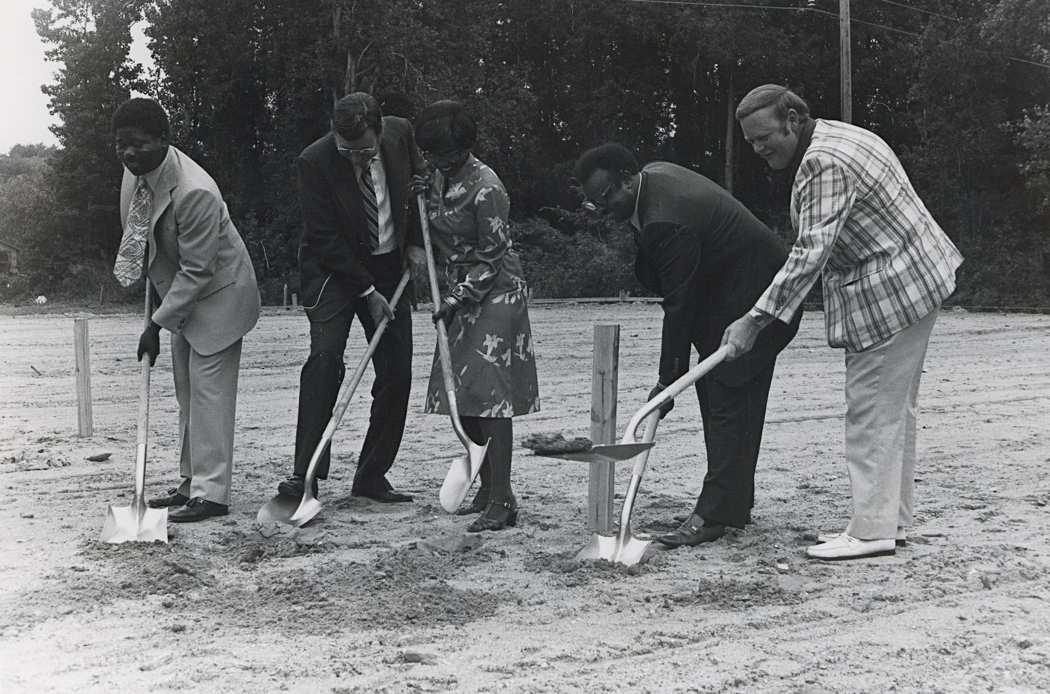 Click to Enlarge Black-and-white photograph of three Black men, a Black woman, and a White man, all well-dressed, shoveling sand with shiny new shovels.