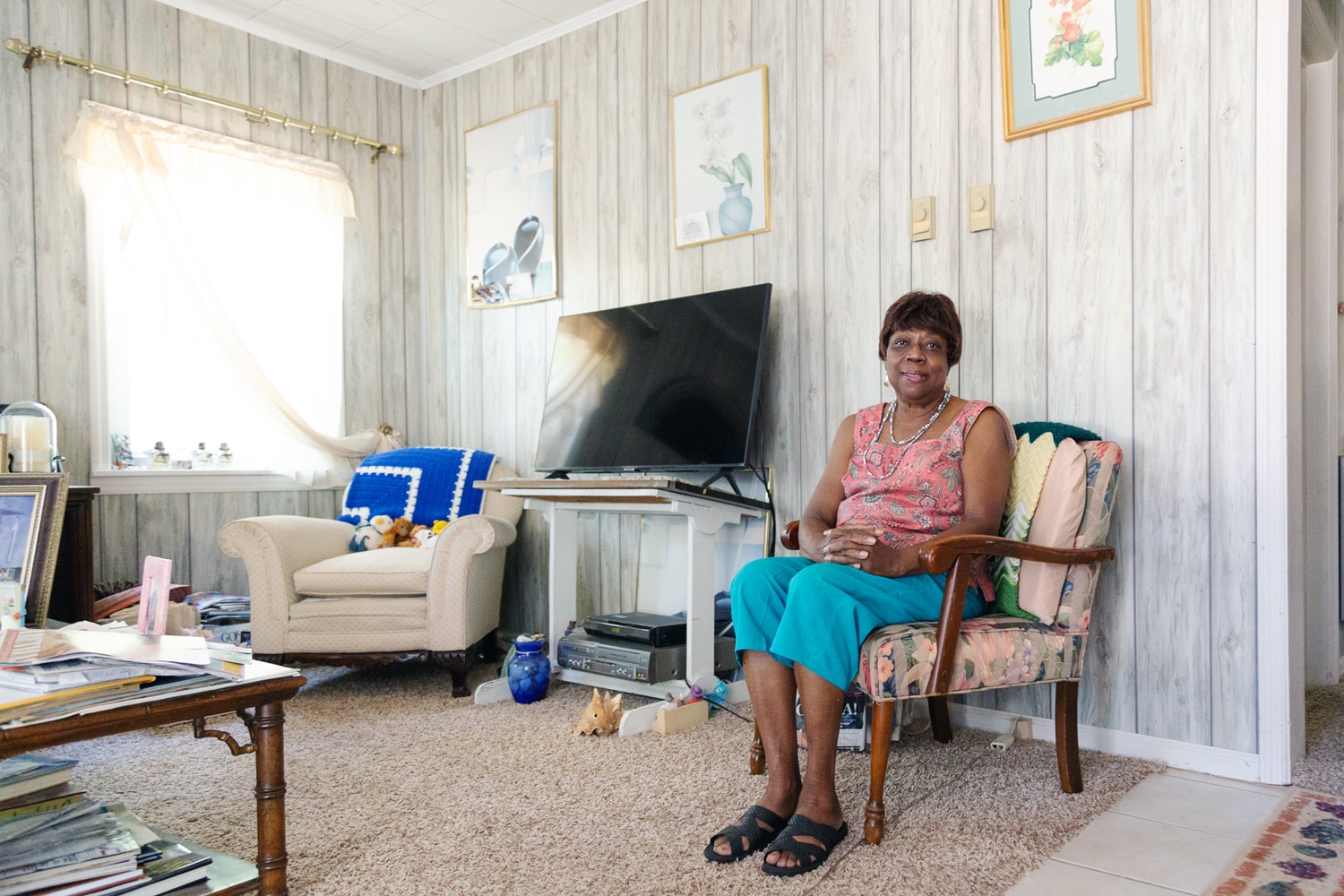 Click to Enlarge Color photograph of middle-aged Black woman in sleeveless blouse, clamdiggers, and sandals, seated in an armchair in her white-paneled living room