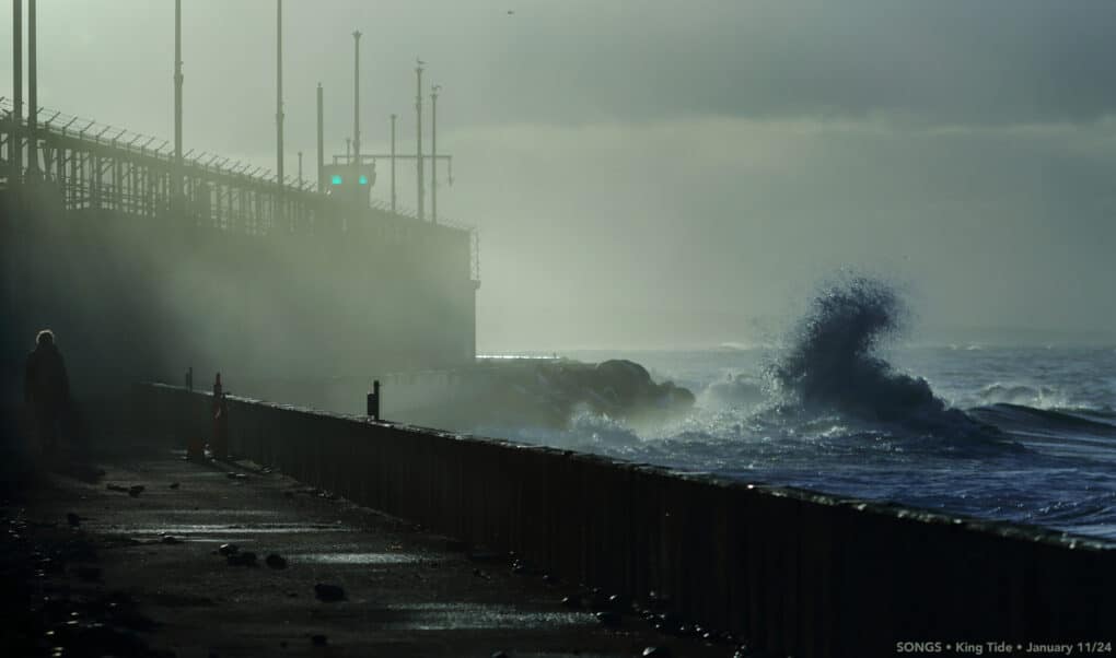 Waves crashing against seawall and water splashing up on walkway with large factory type building behind. 