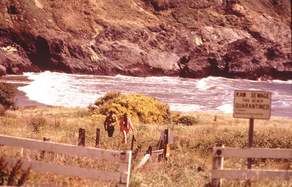Picturesque beach cove, with two walkers and sign in the foreground that says: Raw Sewage. This Beach Quarantined.