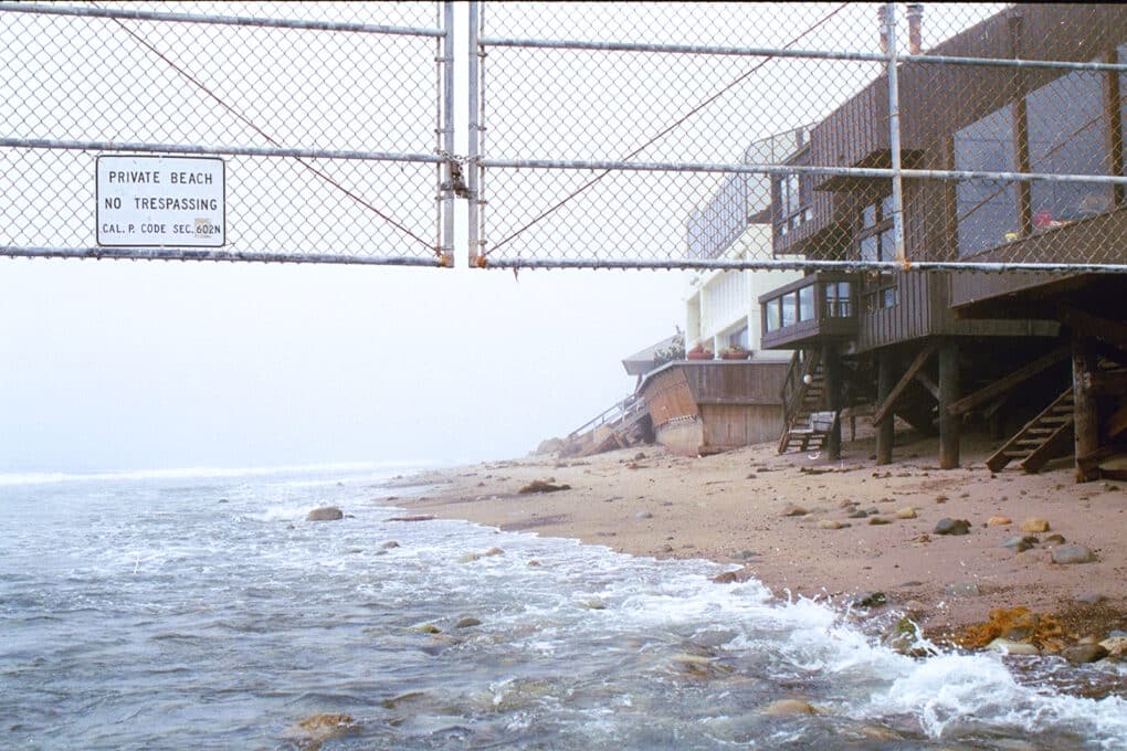 A chain link fence extends over the ocean, with a sign on the fence reading "Private Beach. No Trespassing."