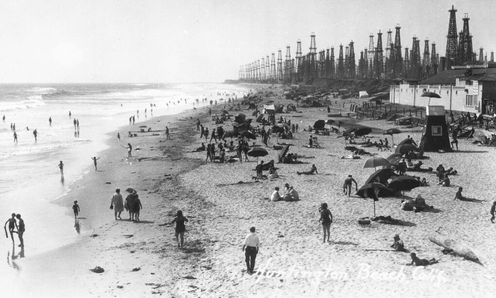 Black and white photo of beach with many people, and in the immediate background, at least 40 tall oil derricks that appear to be nearly on the sand. 