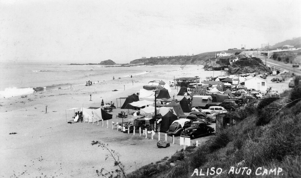 Black and white photo of many cars and tents on the beach. 