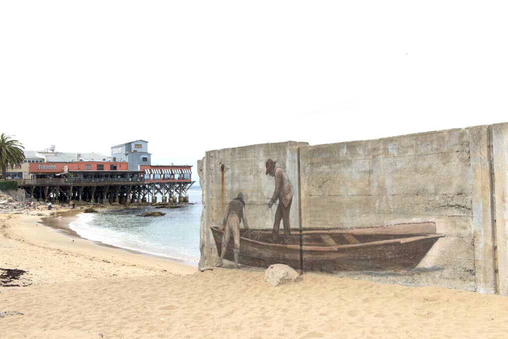 A seawall is painted with a realistic-style image of two men with nets and a fishing boat. 