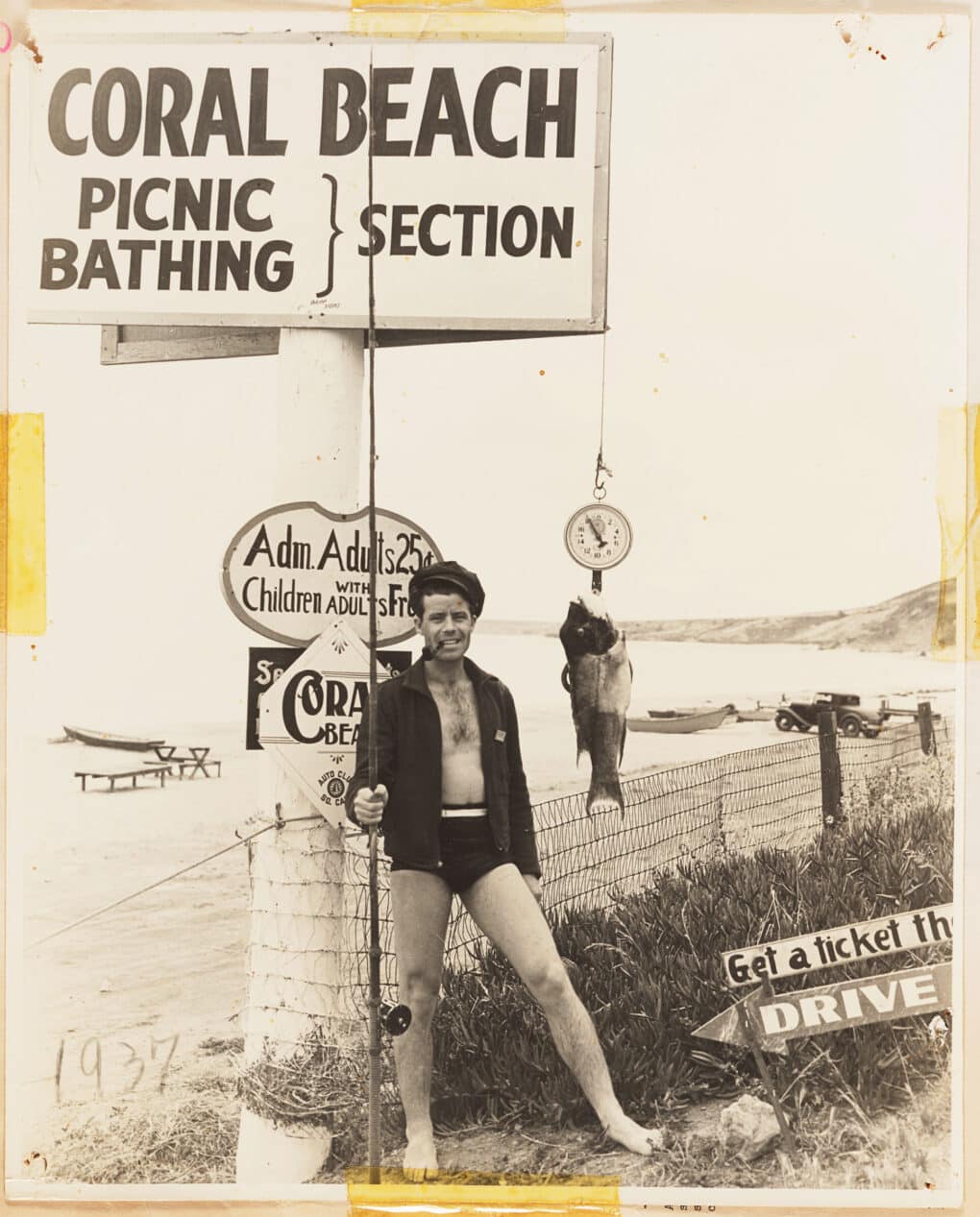 Man is posing in his swimsuit with fishing pole and a large fish. He has a pipe in his mouth and stand directly in front of a sign advertising the beach as a place to picnic and bath, adults cost 25 cents. 