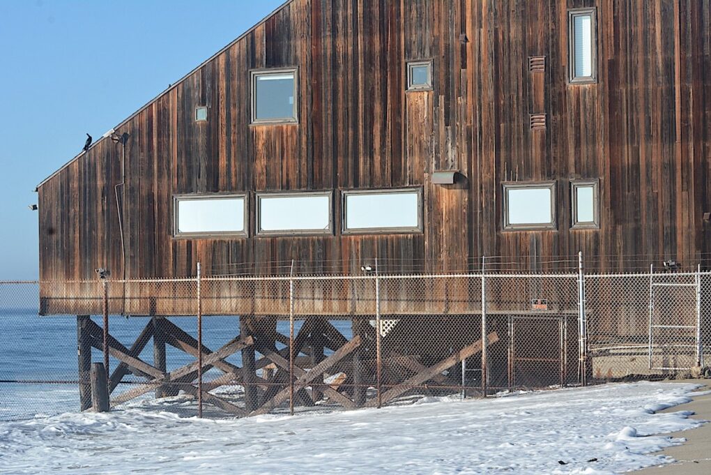 Unusually shaped unpainted wood house is supported by moorings that are entirely in the water. House is behind a chain link fence. 