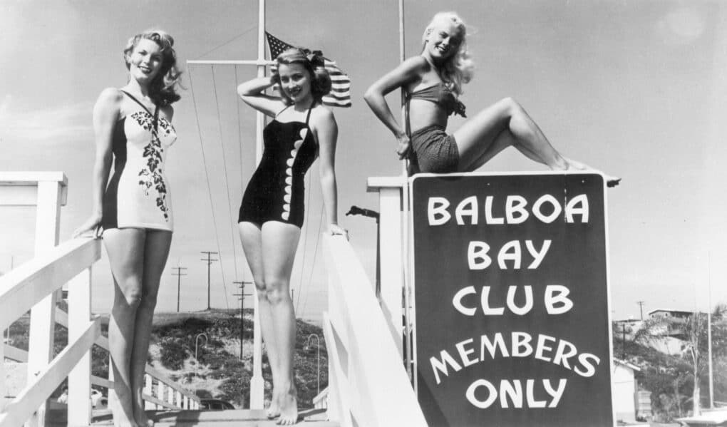 Three pretty White blond girls wearing stylish swimsuits are posing next to a sign that says, "Balboa Bay Club, Members Only."